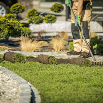 A gardener in tan pants and green gloves uses a rake to lay fresh sod near a stone border. Neatly trimmed shrubs are in the background, conveying an orderly garden scene.